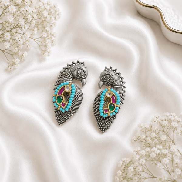 A pair of German silver peacock-shaped statement earrings featuring turquoise beads, green, and magenta pink stones, displayed on a soft white silk fabric background.