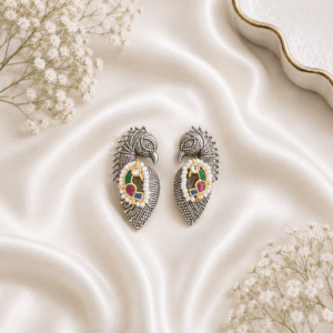 A pair of german silver peacock-shaped statement earrings featuring turquoise beads, green, and magenta pink stones, displayed on a soft white silk fabric background.