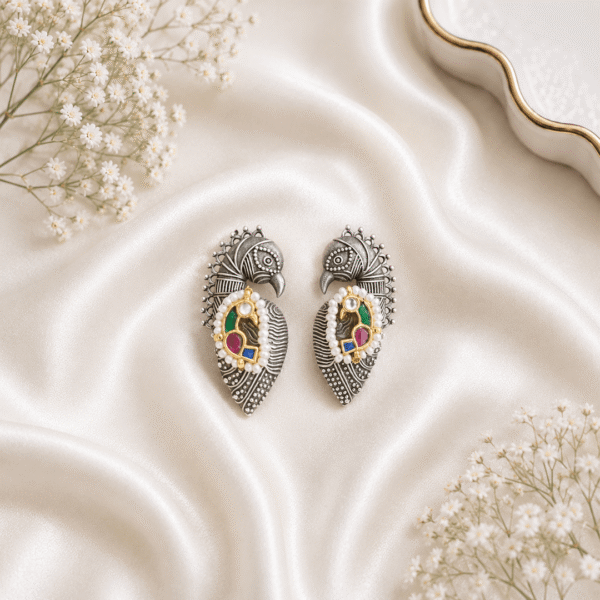 A pair of german silver peacock-shaped statement earrings featuring turquoise beads, green, and magenta pink stones, displayed on a soft white silk fabric background.