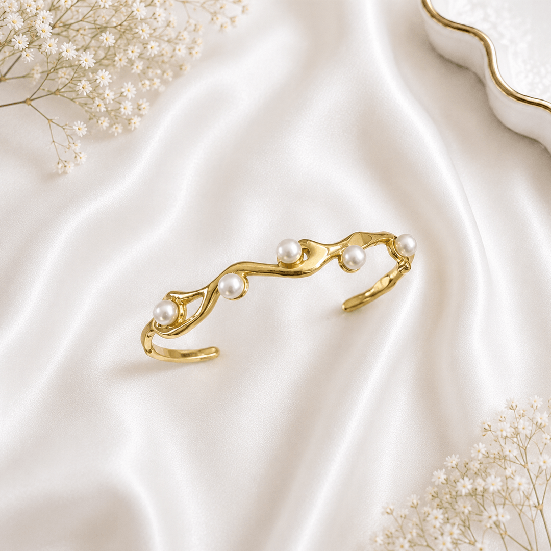 A gold-plated open cuff bracelet featuring an organic, fluid metal design adorned with four lustrous pearls, resting on white satin fabric.