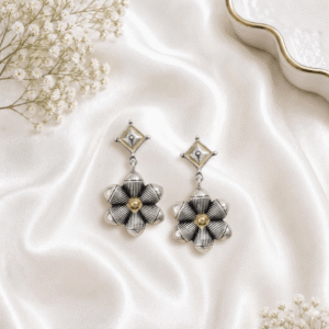 A pair of elegant oxidized silver drop earrings featuring a diamond-shaped stud top and a prominent six-petaled flower design, resting on a luxurious white satin background.