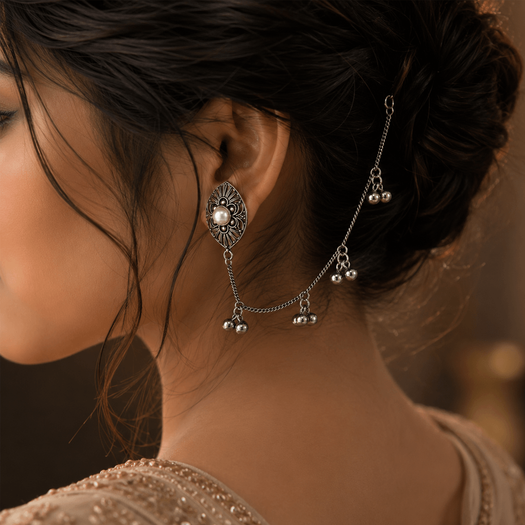A close-up view of a traditional oxidized silver earring featuring an intricate oval motif with a central pearl, connected to an elegant ear chain (kansar) that drapes along the hair and features small metallic dangles.