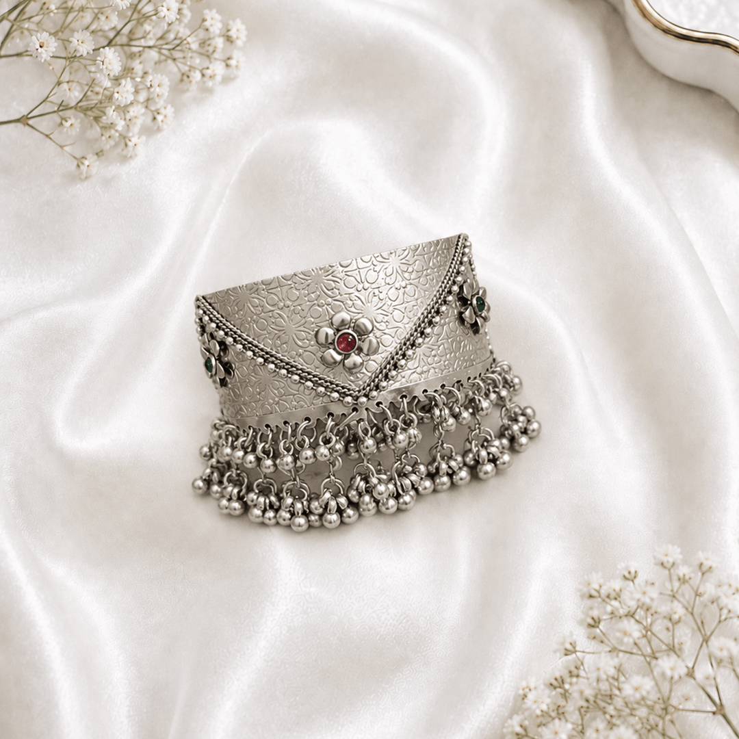 Oxidized silver kada bracelet with floral engraved design, red and green stone accents, and hanging ghungroo bead detailing on white satin background