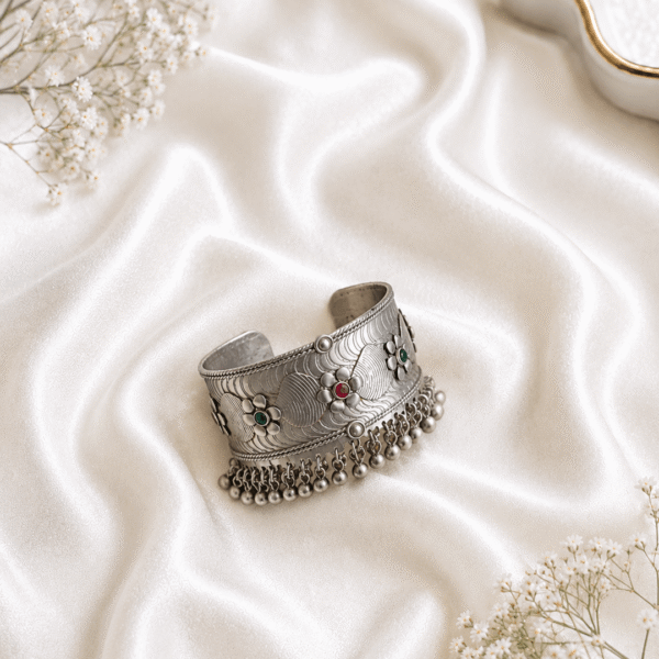 Oxidized silver kada bracelet with floral engraved design, red and green stone accents, and hanging ghungroo beads on white satin background