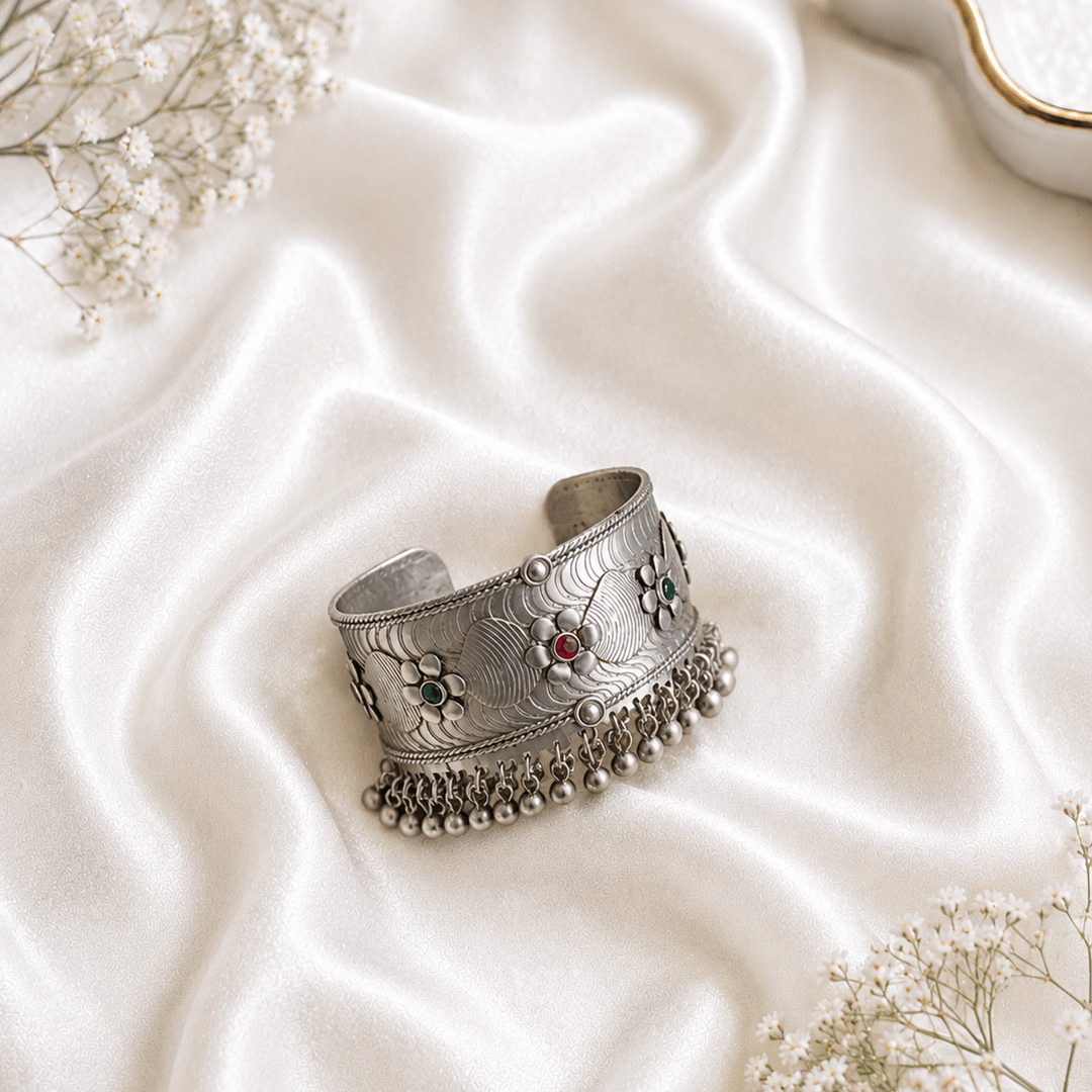 Oxidized silver kada bracelet with floral engraved design, red and green stone accents, and hanging ghungroo beads on white satin background
