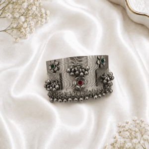 Oxidized silver wide kada bracelet with floral motifs, red and green stone accents, and ghungroo bead detailing on white satin background