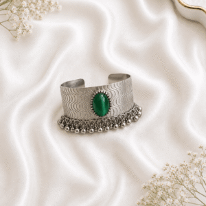 Oxidized silver kada bracelet with green stone and ghungroo bead detailing on white satin background