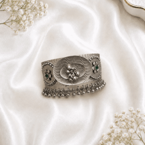 Oxidized silver kada bracelet with oval center motif, green stone floral side design, engraved pattern, and hanging ghungroo beads on white satin background