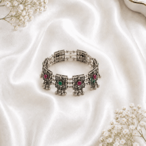 Oxidized silver kada bracelet with square engraved panels, pink and green stones, and ghungroo bead detailing on white satin background