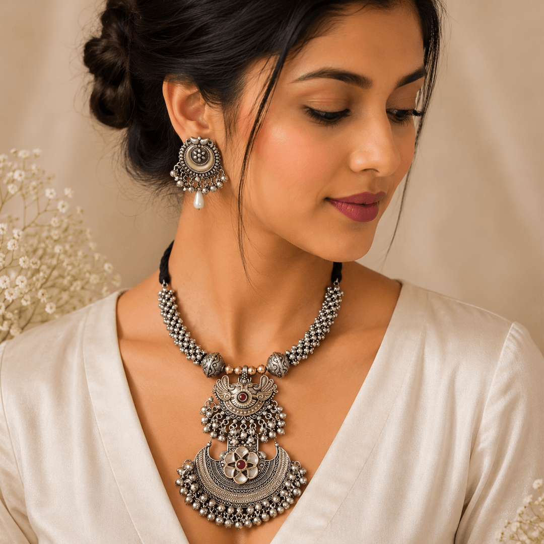 A woman wearing a long, oxidized silver-toned statement necklace with intricate metalwork and stone accents, paired with matching dangle earrings.
