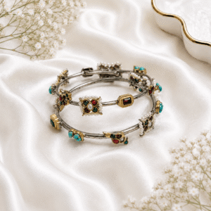 A pair of silver-tone hinged bangles featuring colorful embedded stones (turquoise, red, and green) and delicate pearl detailing, resting on a white satin background.