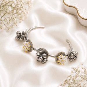 An elegant, open-ended silver-tone kada bracelet featuring alternating clusters of white pearls and metallic ghungroo bells, displayed on a draped white satin fabric.