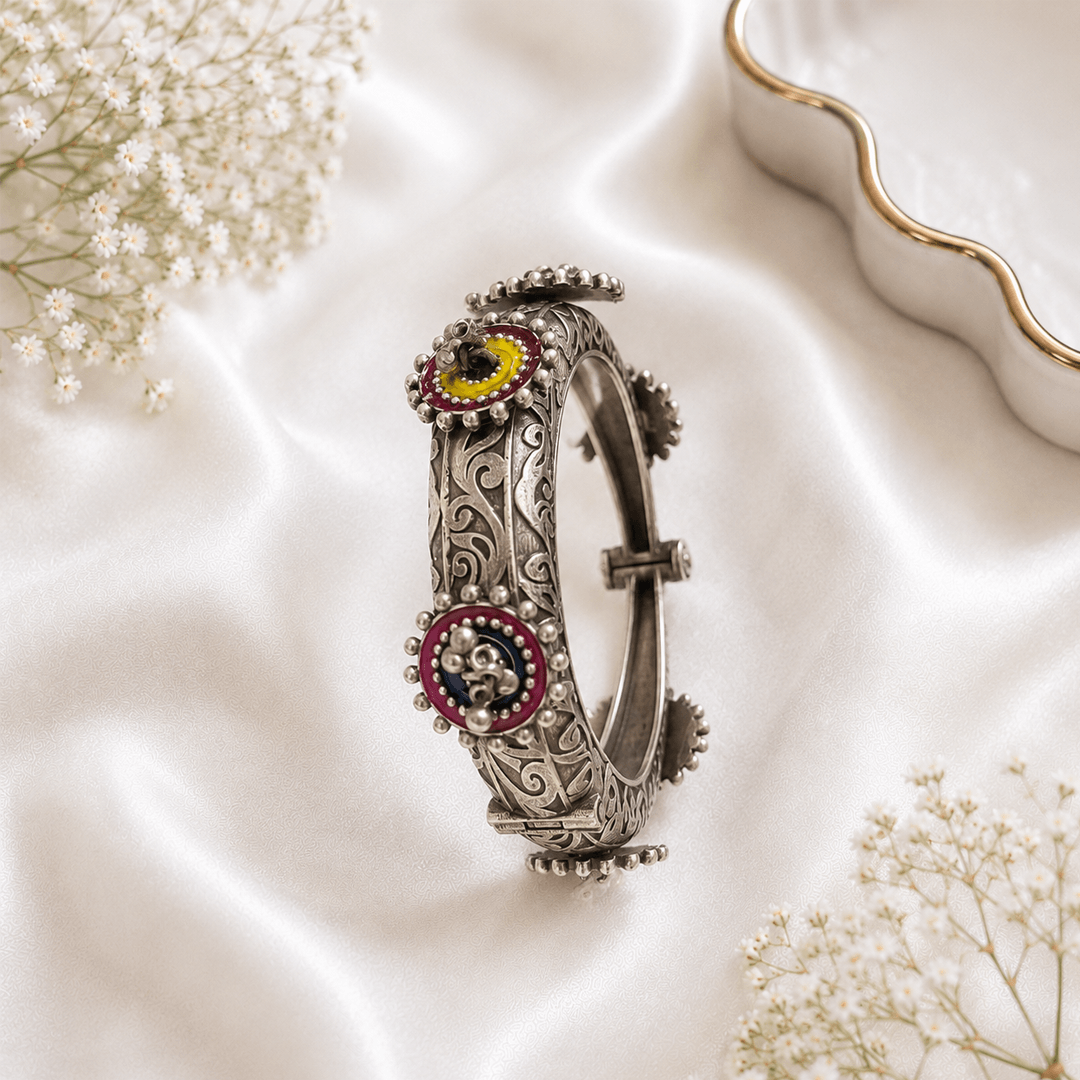 An ornate, printed silver-tone kada featuring intricate swirling engravings and circular stations decorated with maroon and yellow enamel, resting on a white satin background.
