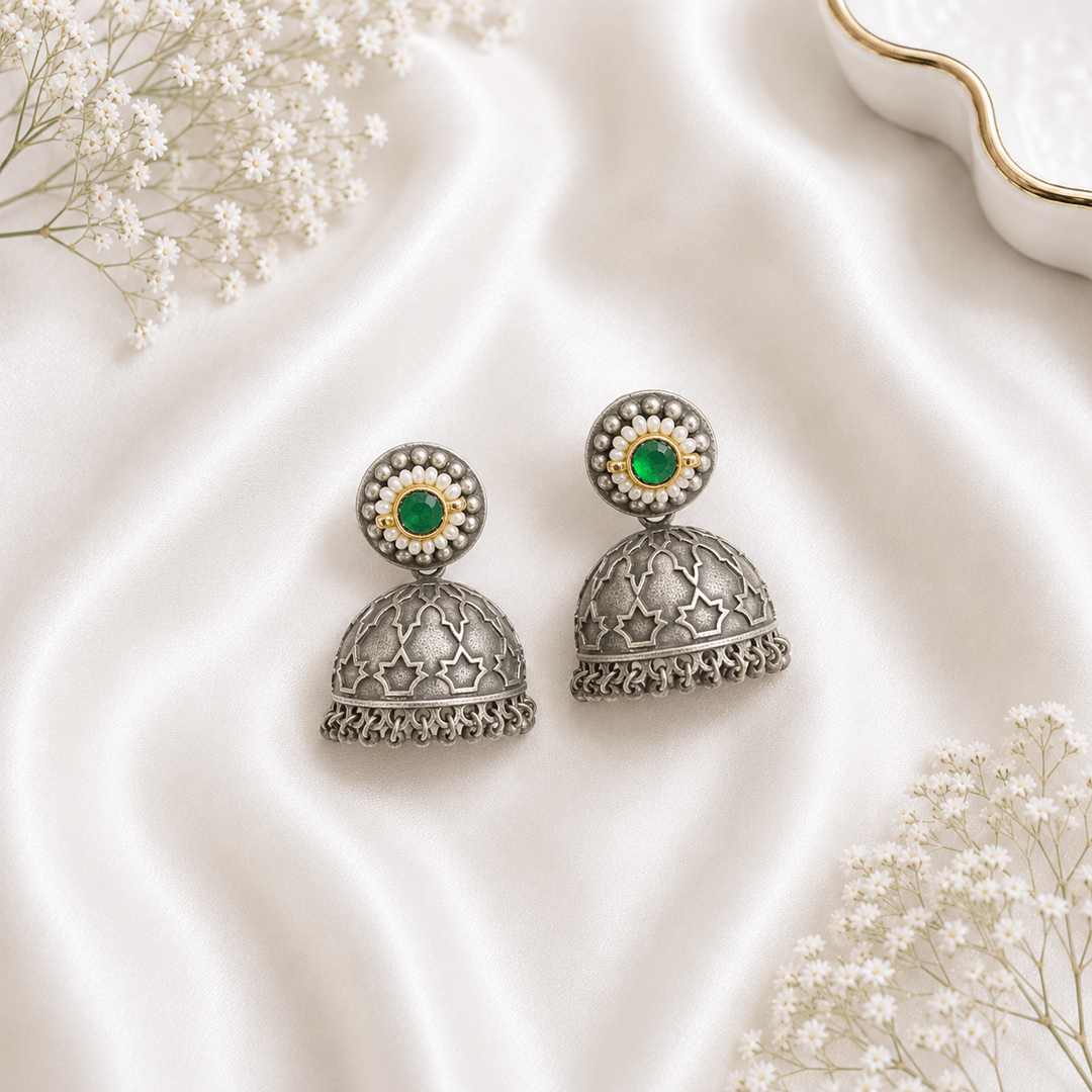 A pair of traditional German silver jhumka earrings featuring a central green stone surrounded by small white pearls, displayed elegantly on white satin fabric with baby’s breath flowers.