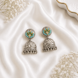 A pair of traditional German silver jhumka earrings featuring a central green stone surrounded by small white pearls, displayed elegantly on white satin fabric with baby’s breath flowers.
