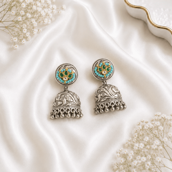 A pair of traditional German silver jhumka earrings featuring a central green stone surrounded by small white pearls, displayed elegantly on white satin fabric with baby’s breath flowers.