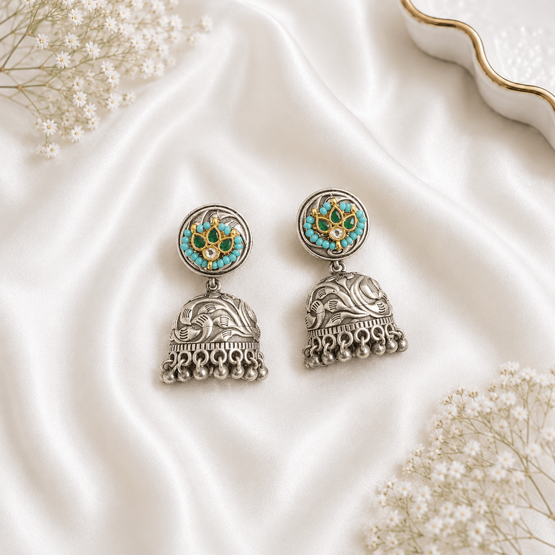 A pair of traditional German silver jhumka earrings featuring a central green stone surrounded by small white pearls, displayed elegantly on white satin fabric with baby’s breath flowers.