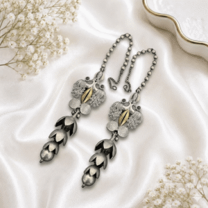 A pair of traditional german silver ear chains (sahare) resting on white satin, featuring intricate peacock motifs with a gold-tone accent, small white stone embellishments, and cascading floral-style metal drapes ending in round beads.
