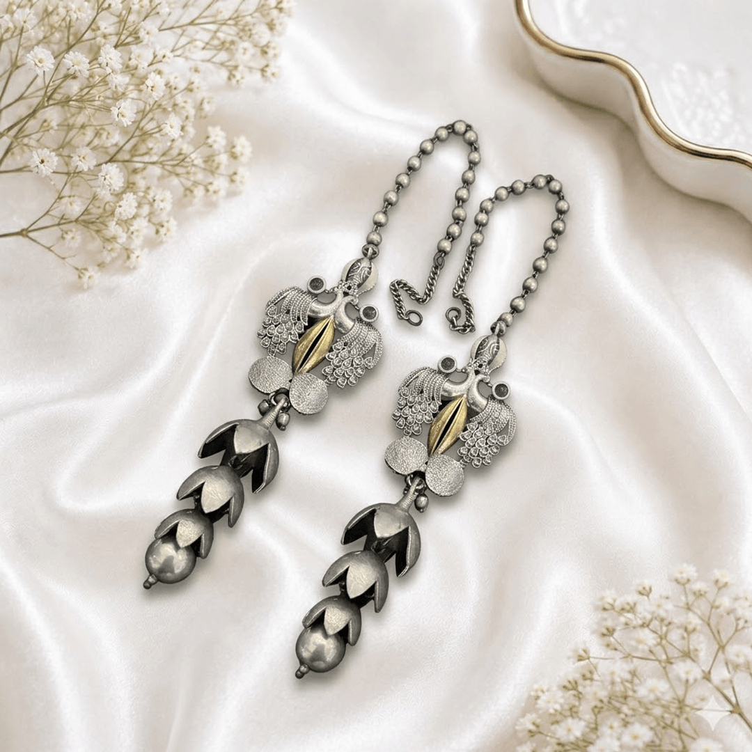 A pair of traditional german silver ear chains (sahare) resting on white satin, featuring intricate peacock motifs with a gold-tone accent, small green stone embellishments, and cascading floral-style metal drapes ending in round beads.