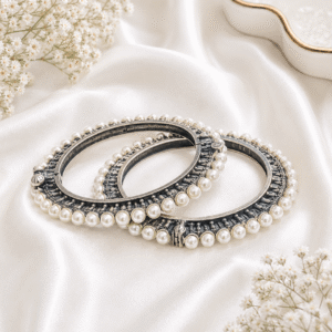 A pair of elegant, hinged german silver bangles featuring a continuous row of white faux pearls along the edges, resting on a draped white satin background.