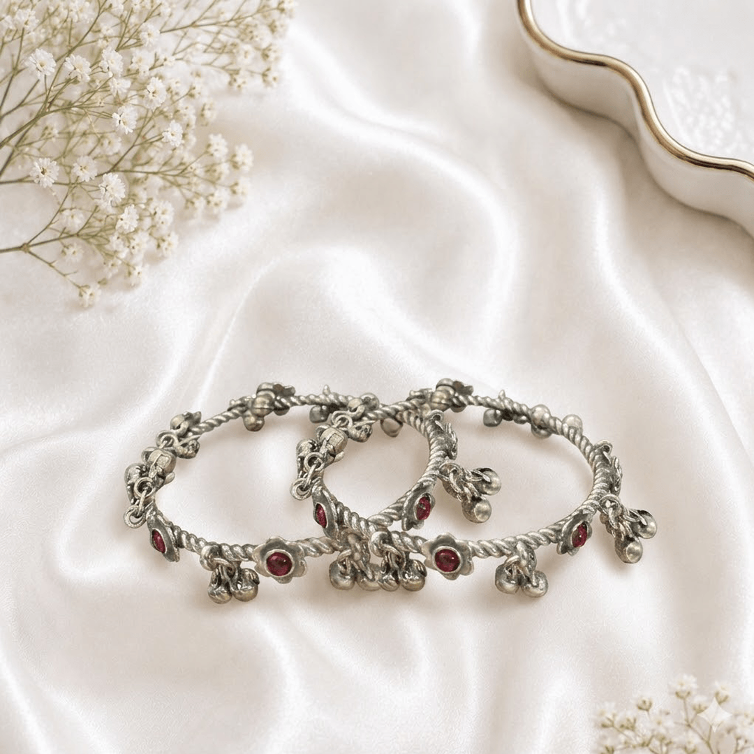 A pair of traditional german silver bangles with a twisted rope design, featuring small floral-set red stones and dangling metal bells (ghungroos), resting on a soft white satin fabric.