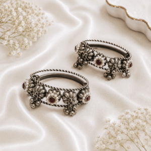 A pair of german silver bangles featuring braided wire detailing, ruby-red stone clusters framed by small white beads, and dangling metallic ghungroo bells, displayed on white satin.