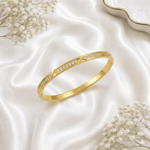 A sleek gold-plated bangle bracelet featuring a recurring geometric "X" pattern interspersed with rows of shimmering clear crystals, resting on a soft white satin fabric.