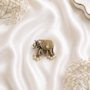 A detailed view of a uniquely designed, german silver and gold-tone ring shaped like an elephant, featuring a prominent white teardrop gemstone and small delicate pearl dangles, resting on a soft white silk fabric background.