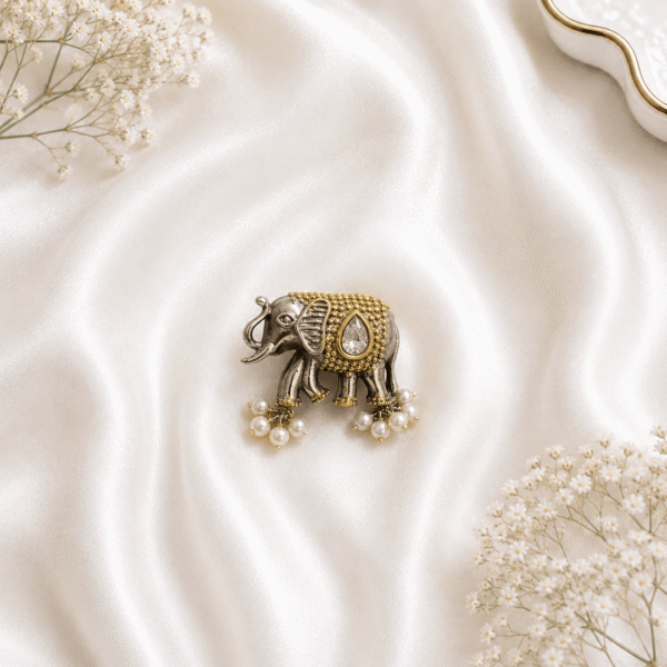 A detailed view of a uniquely designed, german silver and gold-tone ring shaped like an elephant, featuring a prominent white teardrop gemstone and small delicate pearl dangles, resting on a soft white silk fabric background.