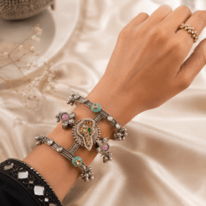 A single, wide, german silver-tone kada bracelet featuring an ornate central section with Kundan stone settings, green and red accents, pearls, and dangling metallic beads, resting on a white satin background.