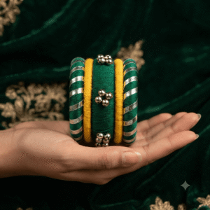 A close-up view of a hand wearing premium green yellow gajara handcrafted thread bangles decorated with colorful cowrie shells, small silver bells, and circular mirror work.
