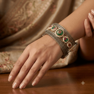 A detailed close-up shot of a handcrafted, antique dual-tone silver and gold kada bangle worn on a woman's wrist. The bangle features intricate Kundan stone work, vibrant green and red Meenakari enamel details, and a filigree pattern, resting on a dark wooden table with a blurred traditional saree in the background.