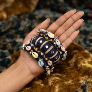 A close-up view of a hand wearing premium navy blue handcrafted thread bangles decorated with colorful cowrie shells, small silver bells, and circular mirror work.