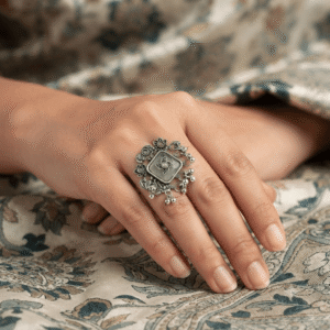 Elegant oxidised silver floral statement ring featuring a square green stone centerpiece with intricate floral detailing and dangling beads, worn on a woman's finger.