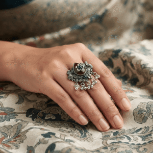 Woman wearing an oxidised silver statement ring with maroon and green stones, floral detailing, and ghungroo charms.