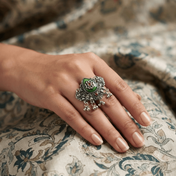 Traditional oxidised silver statement ring featuring a green enamel floral centerpiece with pink stone accents and dangling ghungroo beads, worn on a woman's finger.