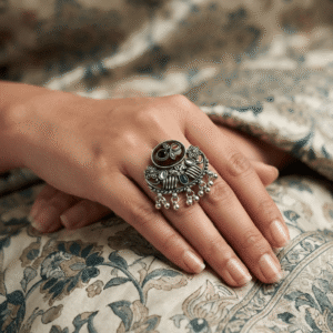 Woman wearing an oxidised silver adjustable statement ring with maroon and green stones, floral detailing, and ghungroo charms.