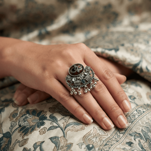Woman wearing an oxidised silver adjustable statement ring with maroon and green stones, floral detailing, and ghungroo charms.