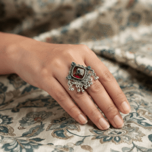 Woman wearing an oxidised silver adjustable statement ring featuring a red square stone, green stone accents, and ghungroo charms.