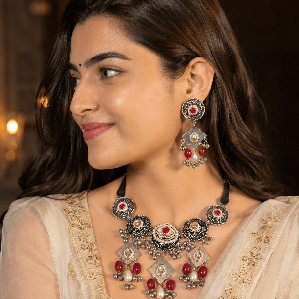 Premium german silver necklace set with ruby red stones, pearl accents, ghungroo detailing, and matching statement earrings worn by a female model.