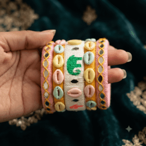A close-up view of a hand wearing premium white pink elephant design handcrafted thread bangles decorated with colorful cowrie shells, small silver bells, and circular mirror work.