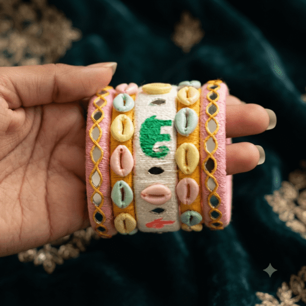 A close-up view of a hand wearing premium white pink elephant design handcrafted thread bangles decorated with colorful cowrie shells, small silver bells, and circular mirror work.