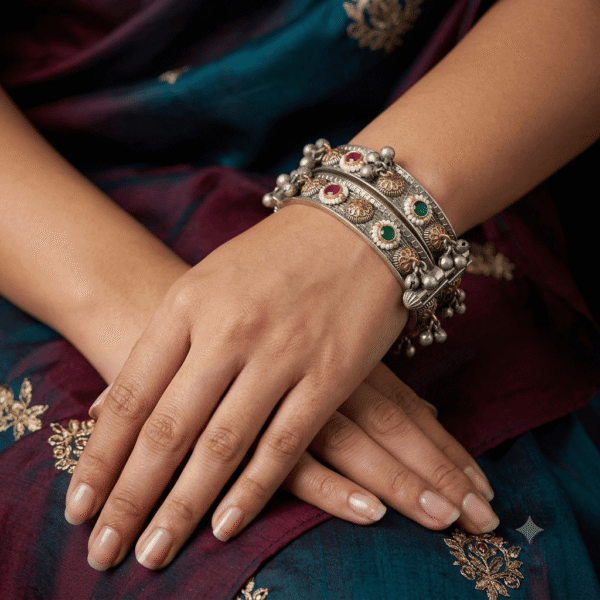 Pair of handcrafted antique dual-tone silver kadas featuring emerald green and ruby red stones encircled with pearl beads, floral gold-tone motifs, and hanging ghungroo cluster drops worn on a wrist.