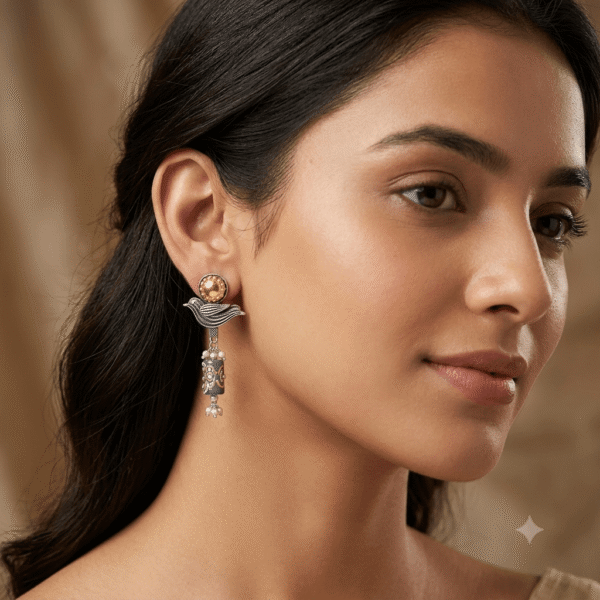 Pair of Kreezo oxidized silver drop earrings featuring a textured golden sun stud, a stylized bird motif, and a cylindrical green enamel bead dangle with delicate white pearls, displayed on a smooth white satin fabric with baby's breath flowers