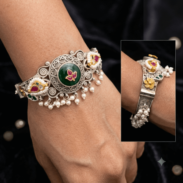 Model wearing an intricate dual-tone oxidized silver cuff bracelet featuring a central round green cabochon with ruby leaf overlay, filigree scrollwork, pearl seed borders, and dangling seed pearl drops.
