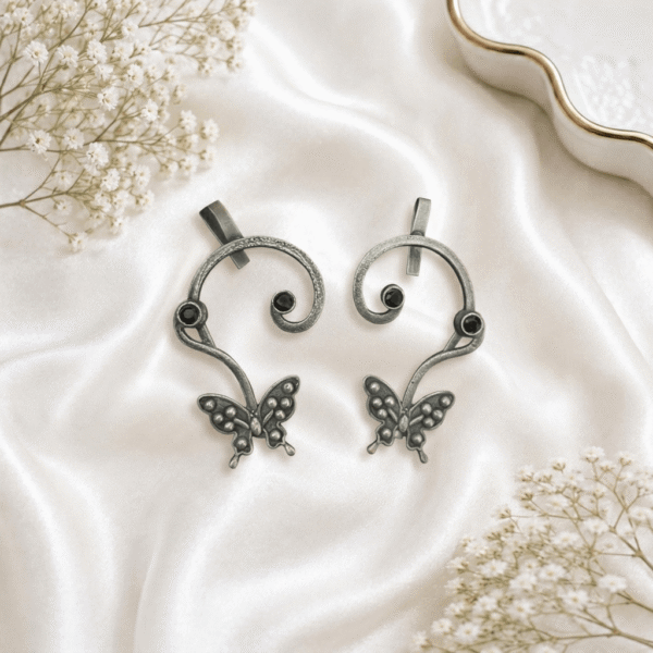 Pair of handcrafted antique oxidized silver ear cuffs featuring swirled vine bands, detailed butterfly bottom charms, and round dark red garnet stone studs, set against a soft white silk background.