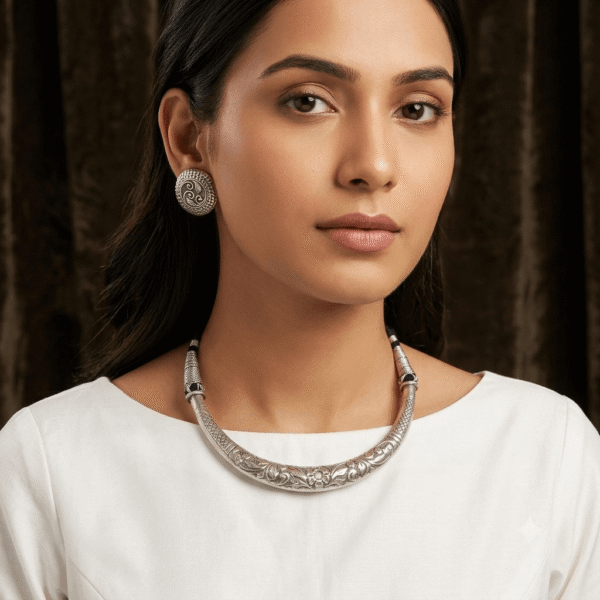 Model wearing a sleek, rigid oxidized silver engraved Hasli choker necklace paired with round filigree swirl stud earrings against a dark textured backdrop.