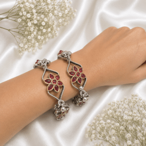 A pair of intricate, floral-inspired german silver bangles featuring ruby-red marquise stones, white pearls, and dangling metallic ghungroo bells, arranged on a white satin background.