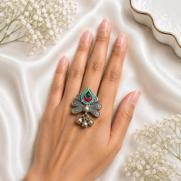 Hand wearing an oxidized German silver statement ring featuring an intricate drop motif with turquoise, green, and ruby stone accents, along with hanging ghungroo bells from Kreezo,