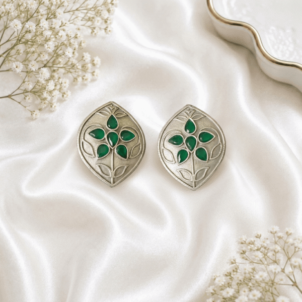 A pair of Kreezo German silver leaf-shaped stud green earrings featuring intricate floral patterns with meenakari stone accents styled on white fabric.